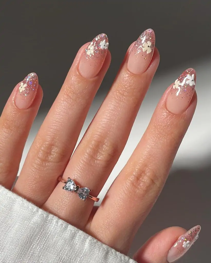 Sparkly ombre nails are two colors, light on the bottom and glittery tips. Flower stickers are small, adhesive designs for a flower look. Together, they create shiny, colorful nails with little flower designs.