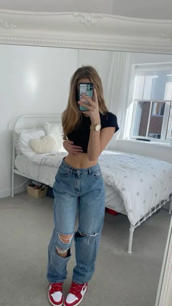 Fashion-forward individual in a cropped black t-shirt, distressed high-waisted jeans, and red sneakers takes a mirror selfie in a bright room.