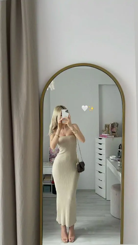 A person stands in front of a mirror wearing a cream ribbed knit dress and holding a shoulder bag.