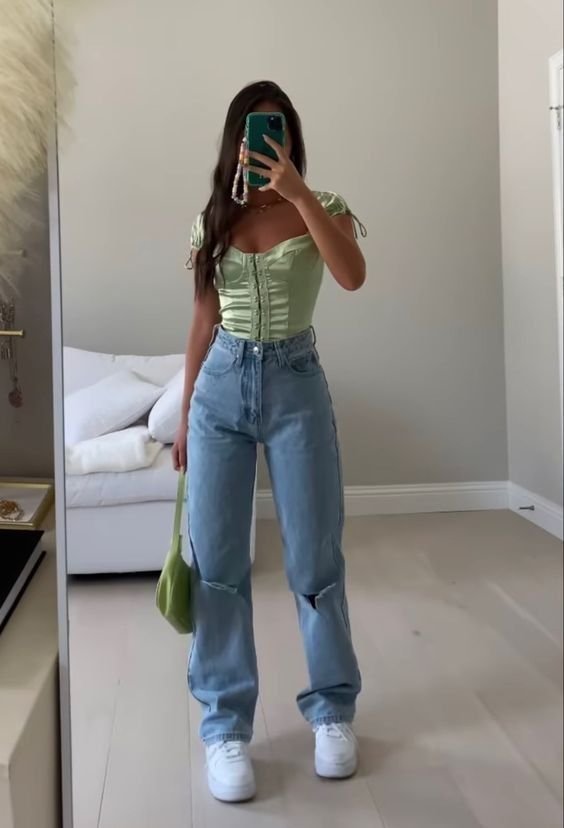 “Fashion-forward individual in high-waisted jeans and green puff-sleeve corset top holding green shoulder bag.