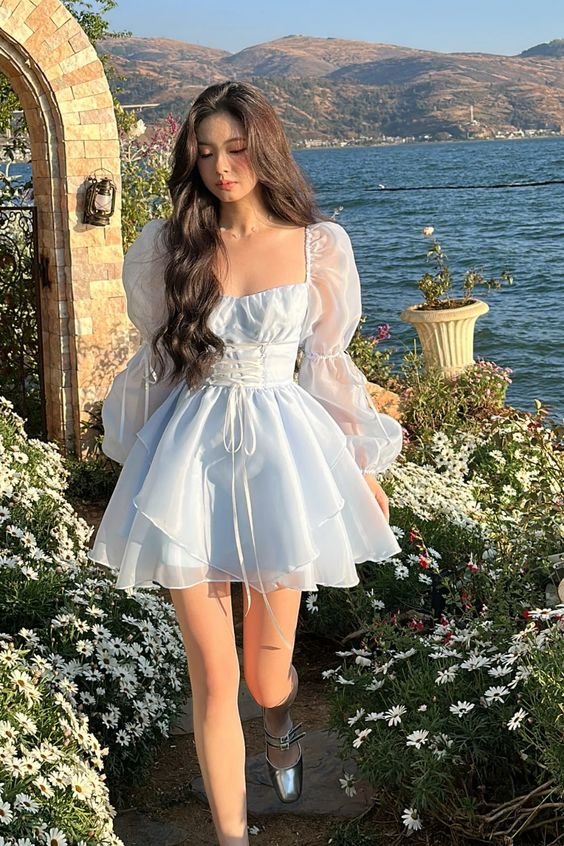 A woman standing by a lake wearing a romantic white puff-sleeve mini dress with sheer details and ribbon tie.