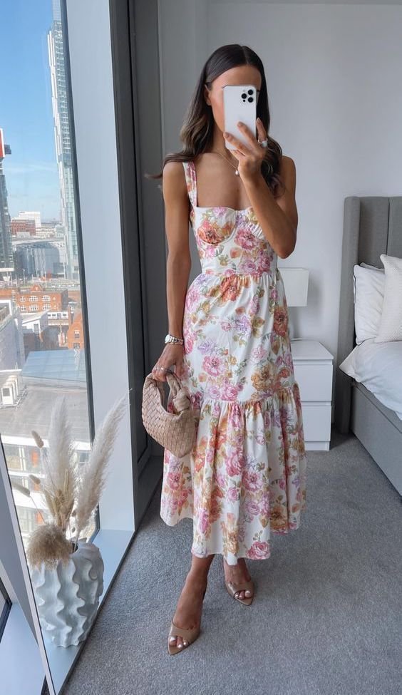 A person in a room wearing a pastel floral dress with tiered ruffles and spaghetti straps, holding a round wicker bag.