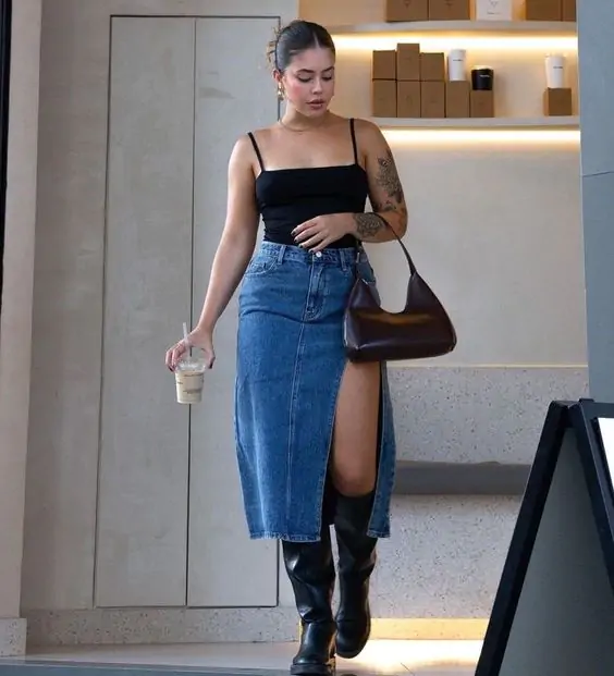 Woman in black top and denim skirt with knee-high boots and brown shoulder bag.