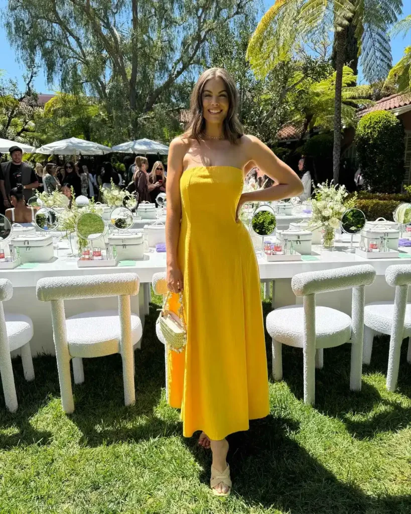 A person at an outdoor event wearing a trendy full-length yellow dress with spaghetti straps paired with nude sandals and carrying a small white floral handbag.