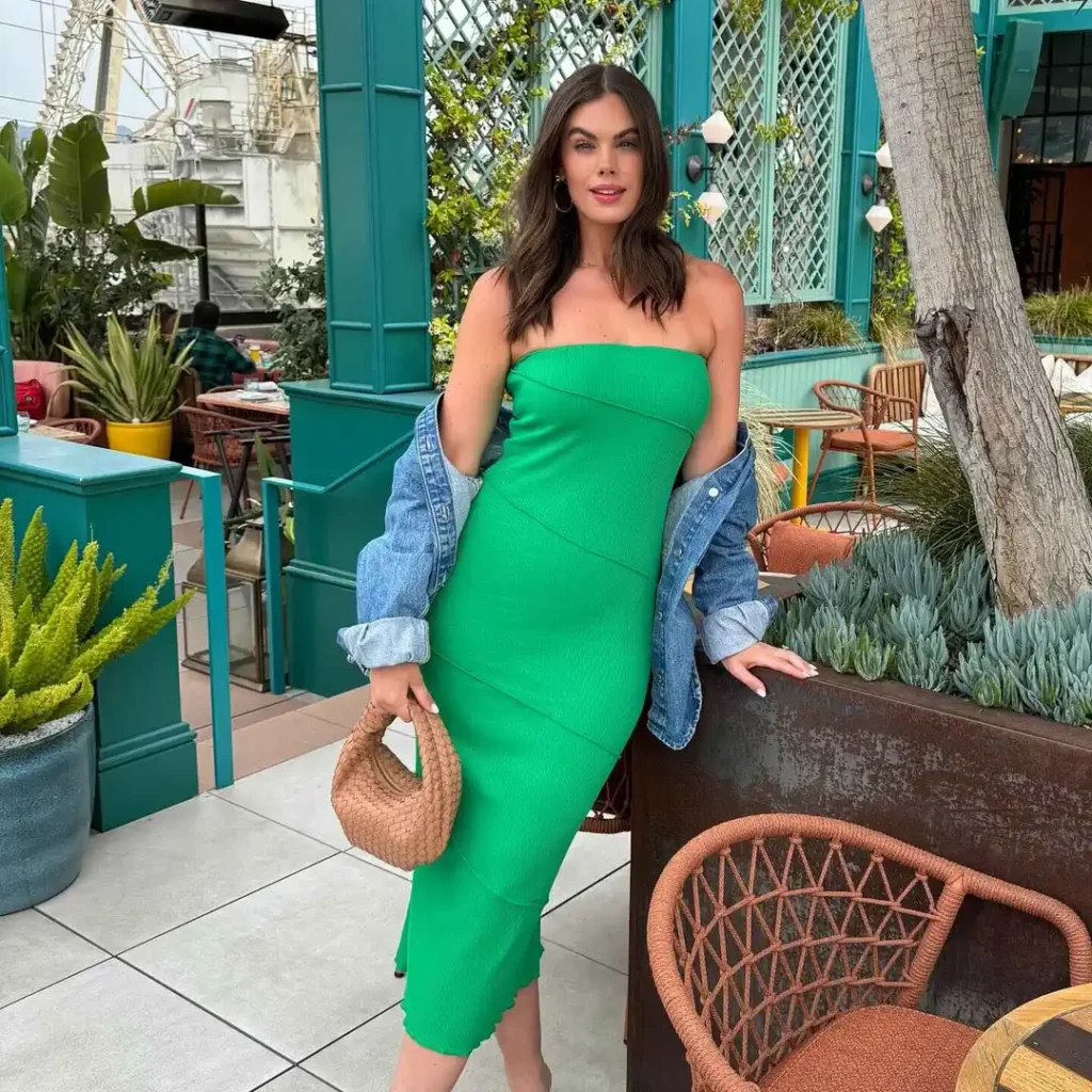 Woman wearing a green strapless midi dress with a blue denim jacket and holding a woven handbag in an outdoor cafe setting.