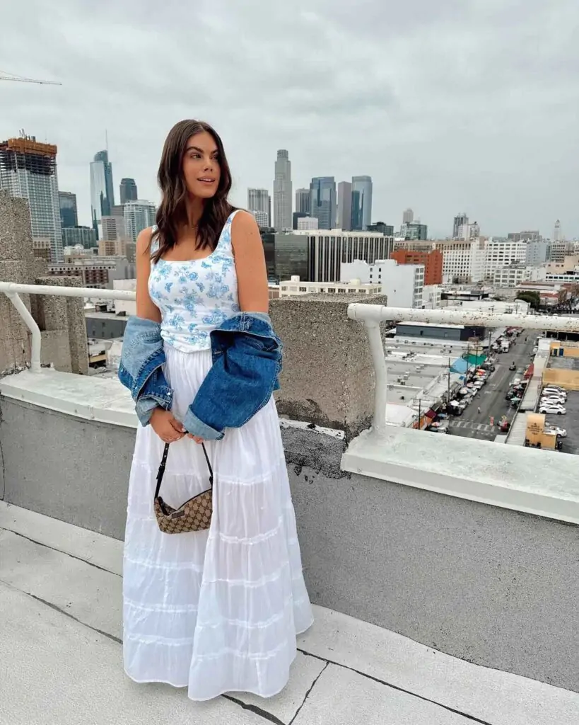 A woman wearing a light blue floral print camisole and a flowing white tiered maxi skirt. She drapes a structured denim jacket over one shoulder and carries a brown leather handbag. The ensemble combines bohemian and urban elements, creating a versatile and stylish look.Bohemian Blend