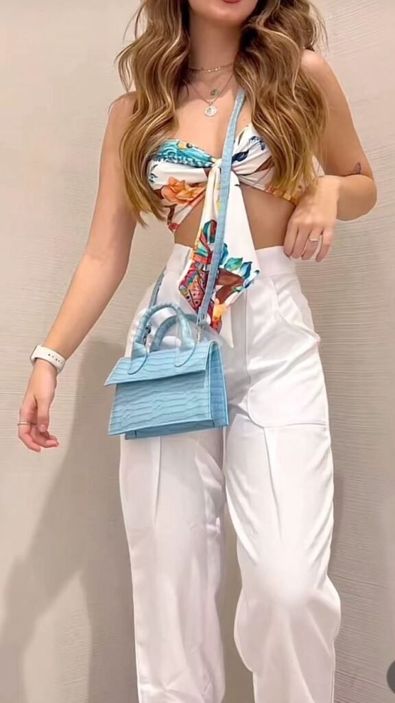 Fashion-forward summer outfit featuring high-waisted white trousers and a tropical print twisted crop top accessorized with a blue quilted handbag.