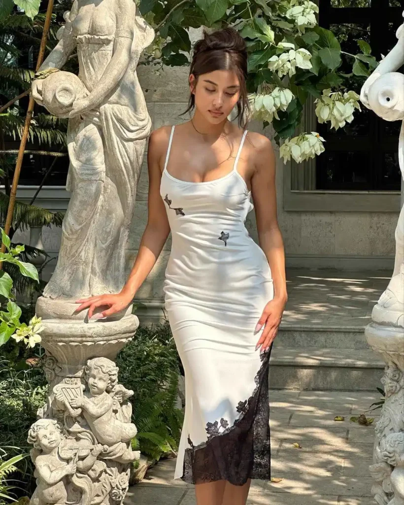 Woman wearing a white slip dress with black lace detailing, standing near a stone statue in a garden