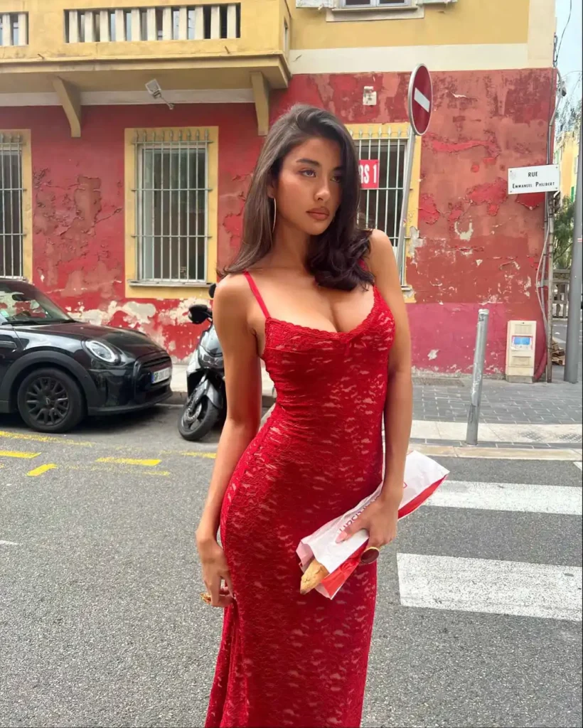 Individual in elegant red floral-patterned full-length dress with thin straps standing on an old city street.