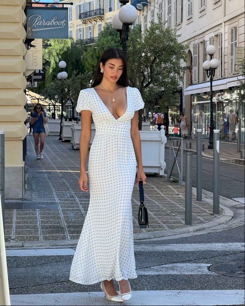 Woman in elegant white polka dot maxi dress with V-neckline and cinched waist paired with white heels on city street.