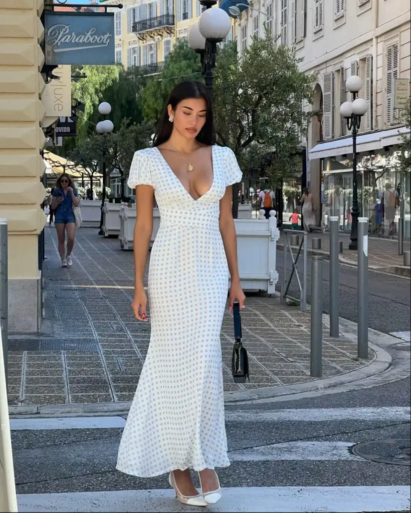 Fashionable individual in white polka-dotted ankle-length dress with V-neckline and short sleeves paired with white heels and black handbag on urban city street.