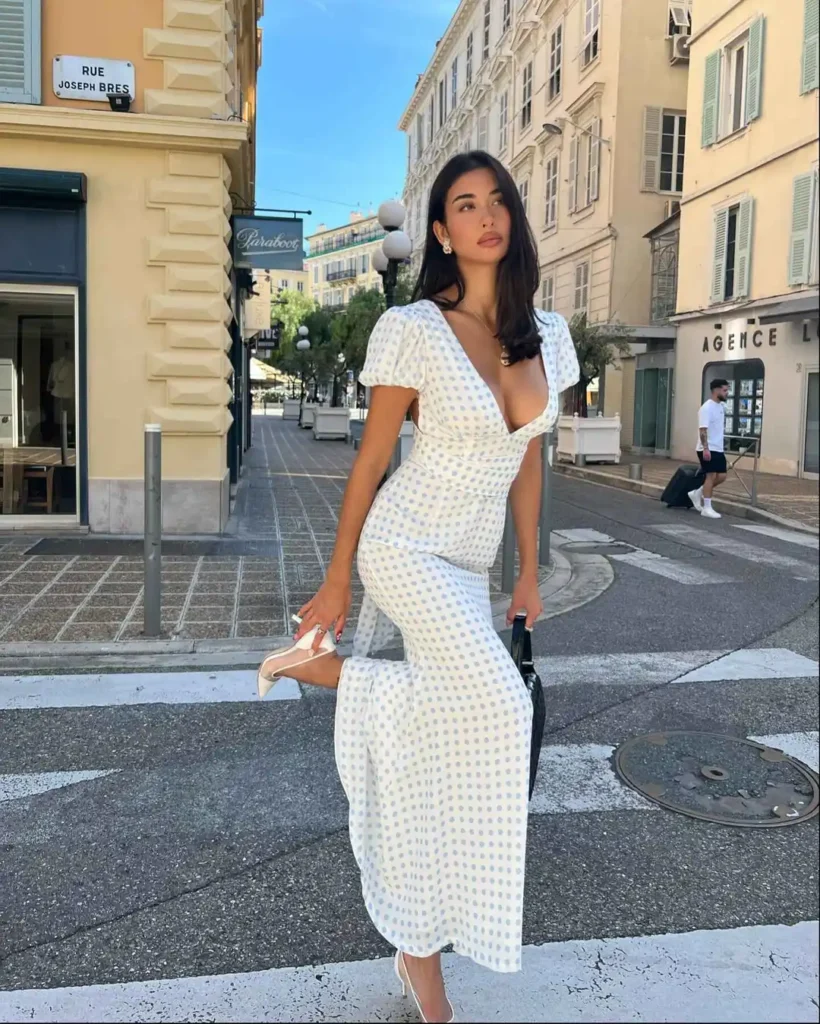 Fashionable individual in white polka-dotted ankle-length dress with V-neckline and short sleeves paired with white heels and black handbag on urban city street.