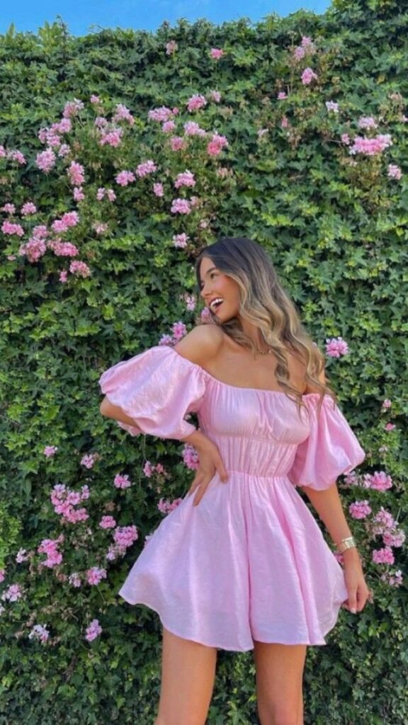 Woman wearing a breezy pink dress with puff sleeves standing against a wall of green ivy and pink flowers.