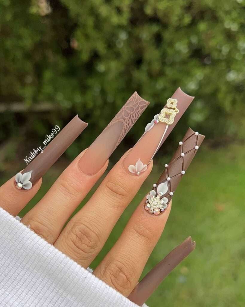 A hand showcasing long, almond-shaped nails painted in earthy tones of rich browns and soft beiges, adorned with delicate white flowers and sparkling gemstones, capturing the essence of autumn.