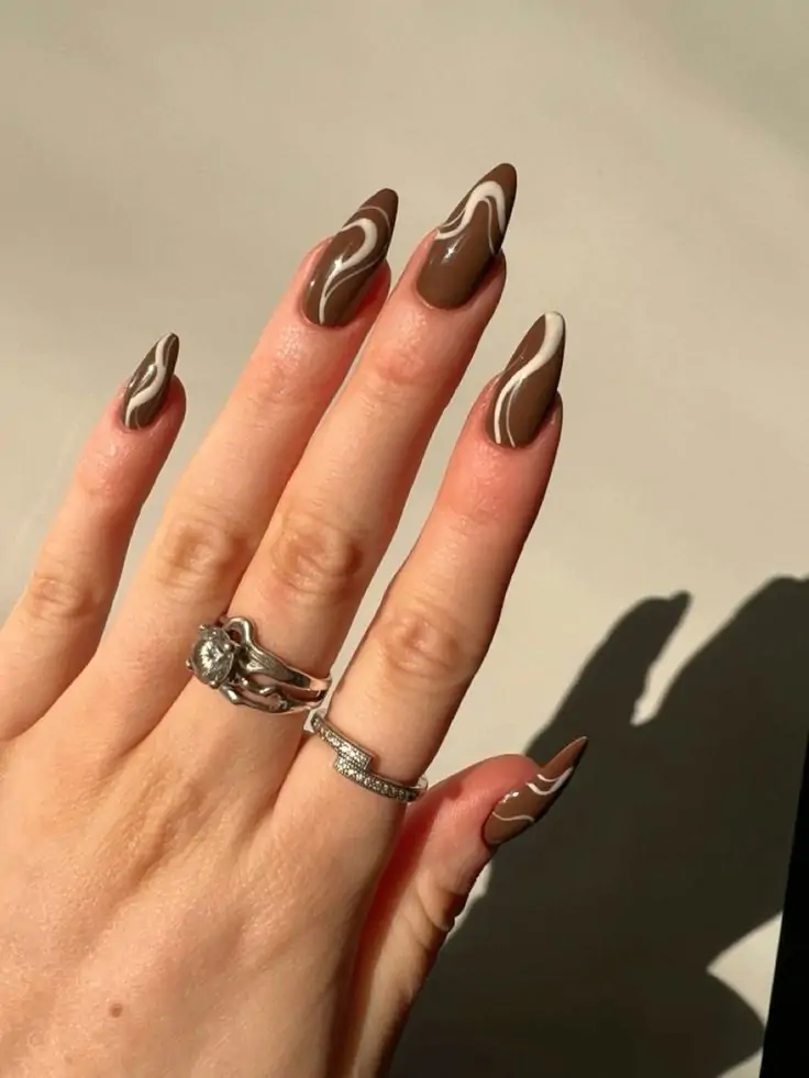 Hand with long, almond-shaped nails painted in fall colors with a glossy finish and accessorized with rings.