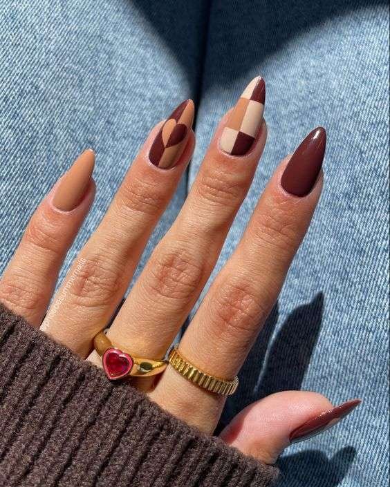 Hand with checkered brown and beige nails, gold ring with red heart stone, on denim fabric.