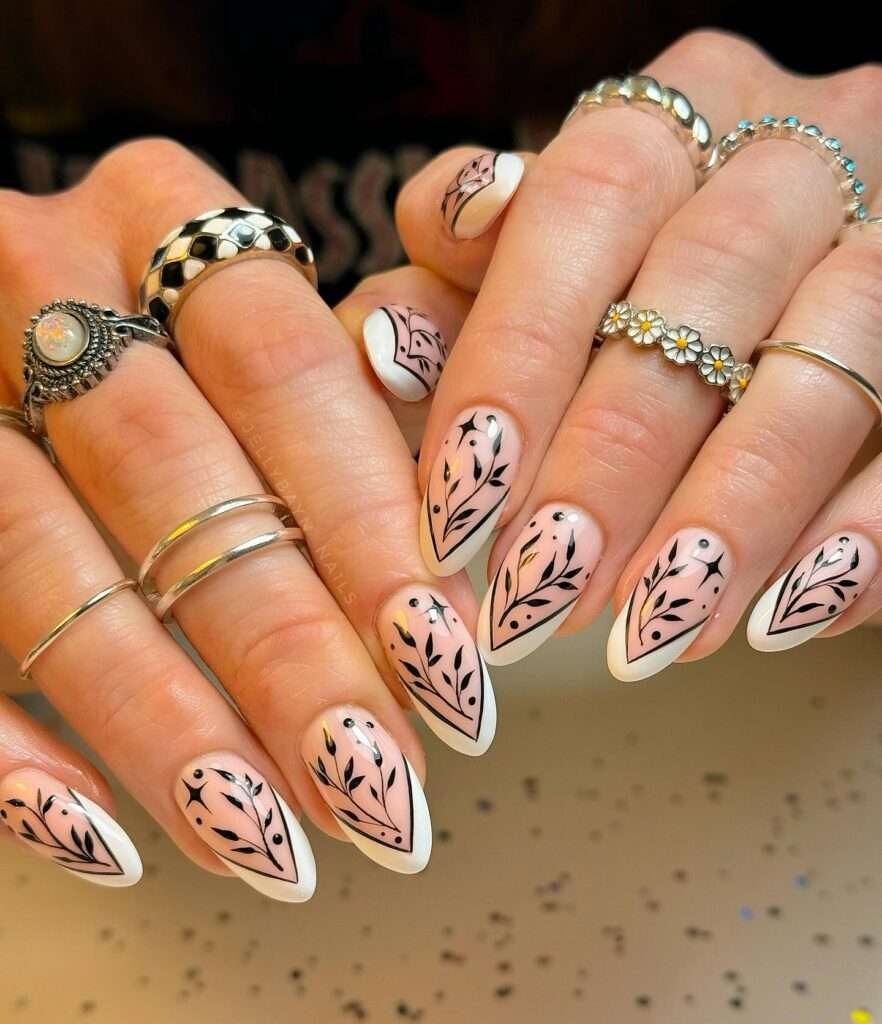 Sophisticated fall nail design with creamy base and intricate black leaf patterns, accessorized with stylish rings.