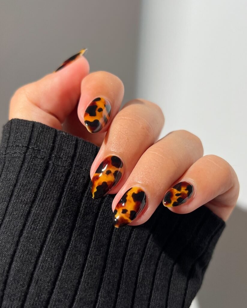 Close-up of almond-shaped nails with leopard print in warm autumn shades against a dark grey sweater