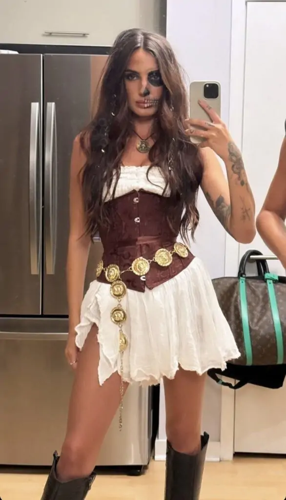 Woman in a pirate-inspired Halloween costume with a brown corset, white dress, gold medallion belt, and skull makeup.