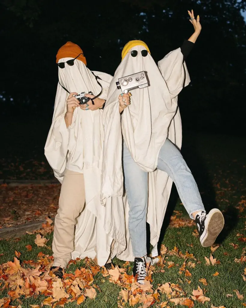  Two people in ghost Halloween costumes with sunglasses and hats posing playfully outdoors at night amidst autumn leaves, one holding a vintage camera.