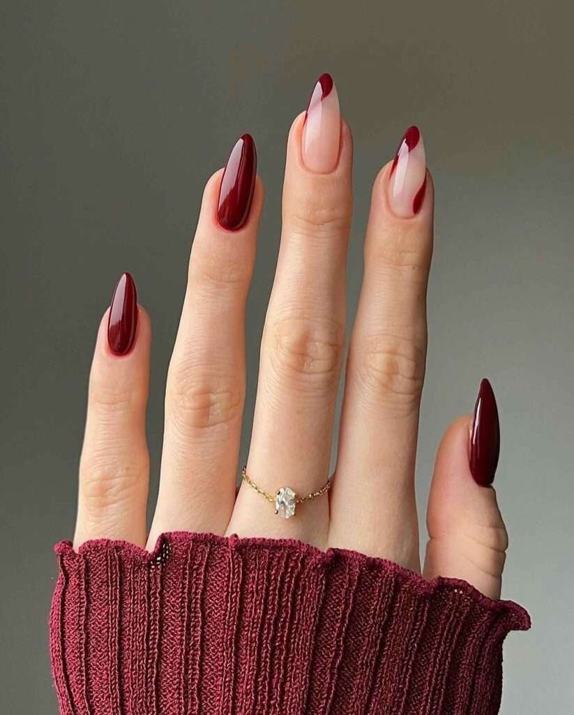 Elegant fall nail design with glossy burgundy nails and white V-shaped accents on ring fingers, complemented by a gold ring and burgundy sweater cuff.