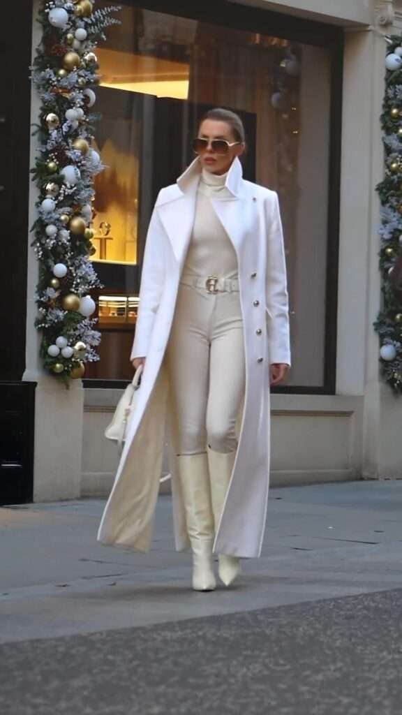  Woman wearing a white long coat with a turtleneck, high-waisted pants, knee-high boots, and a mini handbag.