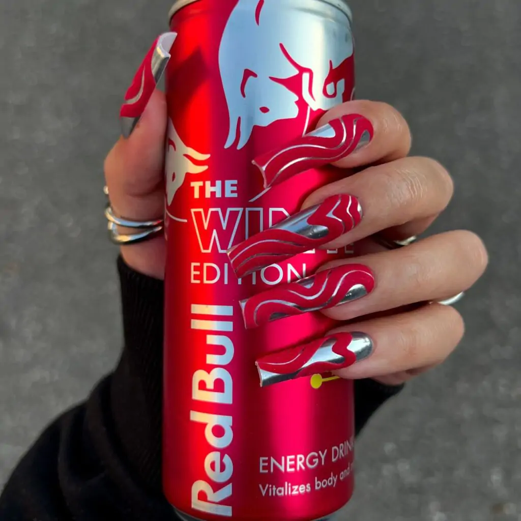 The image shows a hand holding a Red Bull Winter Edition energy drink can. The nails, long and elegant, feature a winter-themed design with red and silver swirls that match the can’s colors. The intricate nail art complements the can's design, making it visually appealing and cohesive.