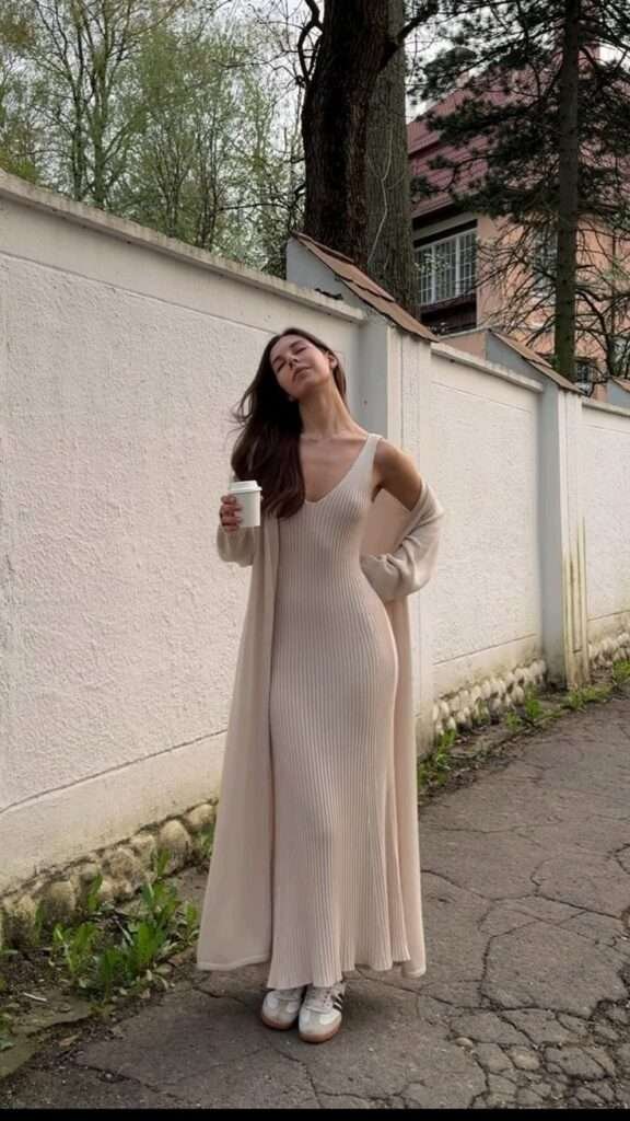 A long, ribbed, beige dress with a fitted silhouette and a scoop neckline. A matching beige cardigan draped over the shoulders, adding a layer of warmth and style. White sneakers with beige accents, providing a comfortable and casual touch.