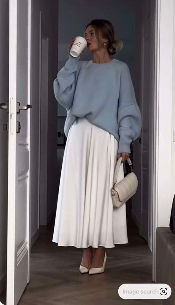 This effortlessly elegant outfit combines a cozy pastel blue oversized sweater with a flowing white pleated maxi skirt. Perfectly styled with pointed white heels and a chic cream handbag, it's a soft and refined look for transitioning into spring.