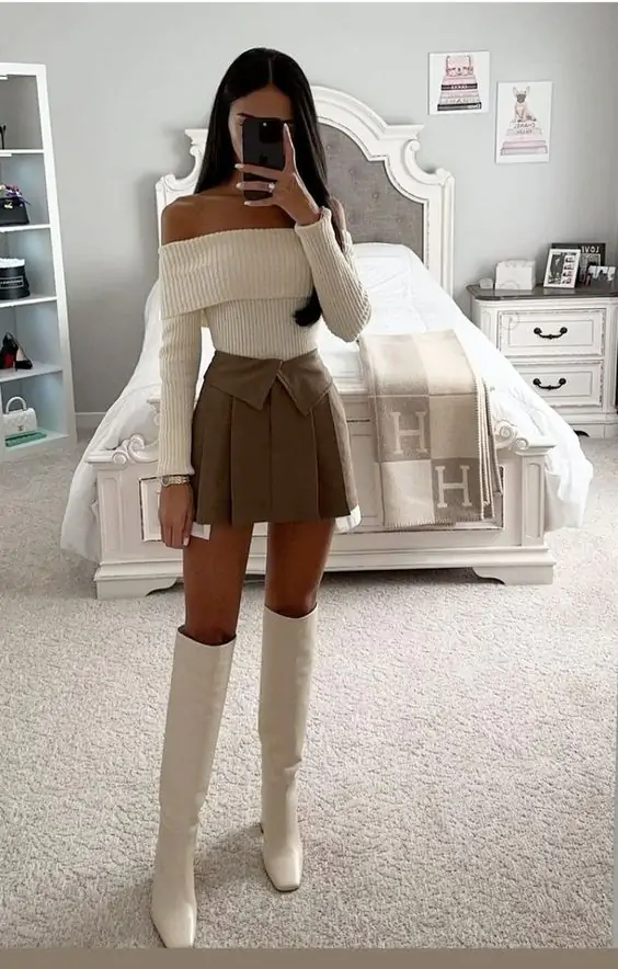 The outfit features a cozy off-the-shoulder sweater paired with a stylish pleated skirt and knee-high boots, perfect for a chic winter look.