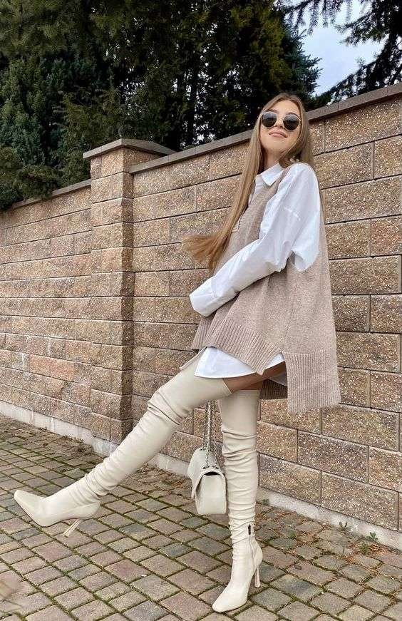 This outfit combines a classic oversized white button-down shirt with a beige sleeveless sweater vest, creating a layered look that’s stylish and cozy. Paired with over-the-knee cream boots, it’s perfect for a warm yet trendy winter vibe.