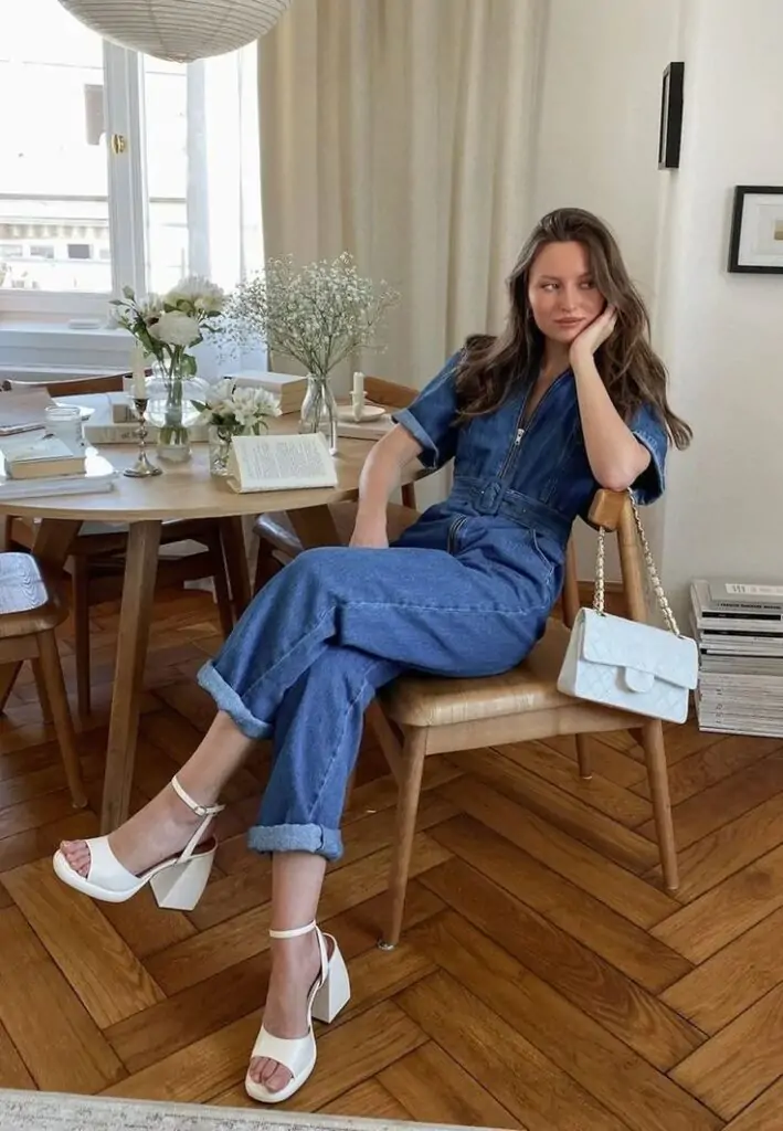 The spring outfit features a trendy denim jumpsuit, perfect for a light and casual yet chic look. It is paired with stylish white high-heeled sandals that add a touch of elegance to the ensemble. The outfit is completed with a sophisticated white quilted handbag with a gold chain strap, adding a pop of refinement to the spring look.