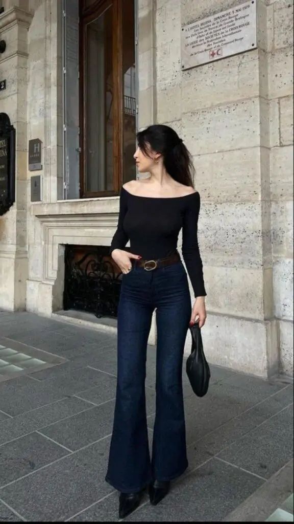 Effortlessly chic, this spring outfit features a black off-the-shoulder top paired with dark blue flared jeans and a brown belt. Black pointed shoes and a small black handbag add a touch of elegance. Perfect for a stylish day out in the city.