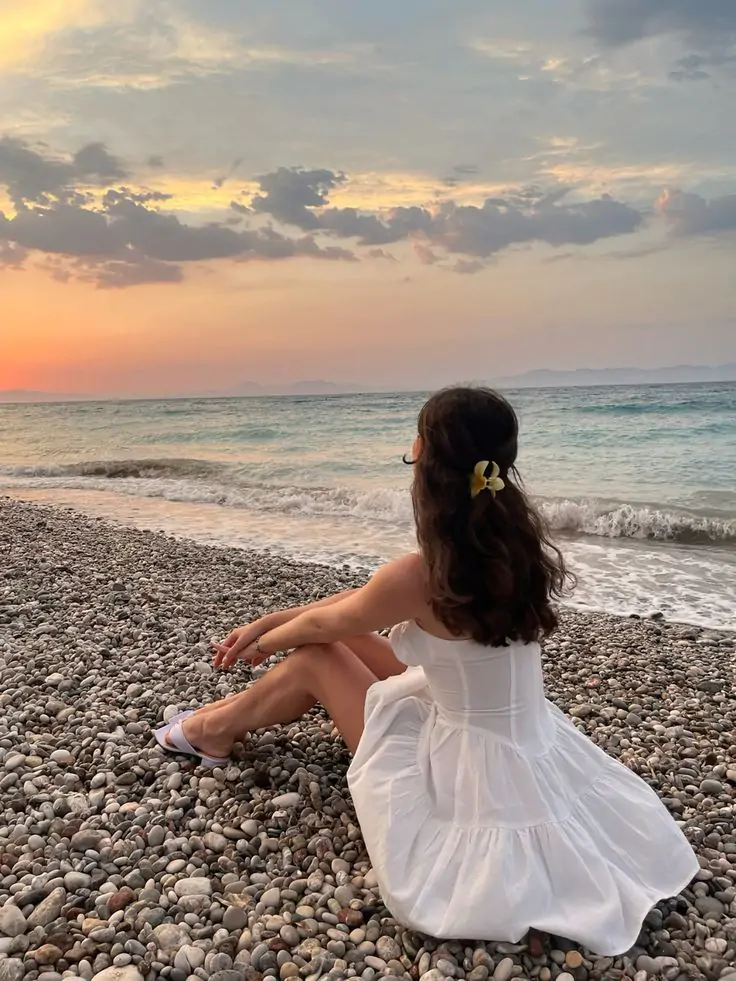 Experience the tranquil beauty of a sunset stroll along a pebbled beach." A timeless white dress flows effortlessly, while a delicate yellow flower hair accessory adds a subtle touch of nature’s elegance. This serene look is perfect for soaking in the peaceful magic of an evening by the sea.