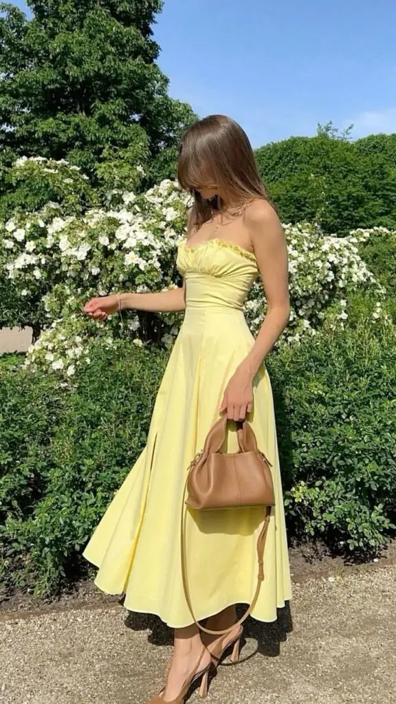 Imagine enjoying a sunny spring day, feeling light and carefree. You're wearing a lovely light yellow, strapless, mid-length dress with a fitted bodice and a flared skirt, capturing the essence of spring. A brown handbag and matching brown high-heeled shoes add a touch of sophistication. Surrounded by lush green foliage and white flowers, this outfit is perfect for a serene, picturesque setting.