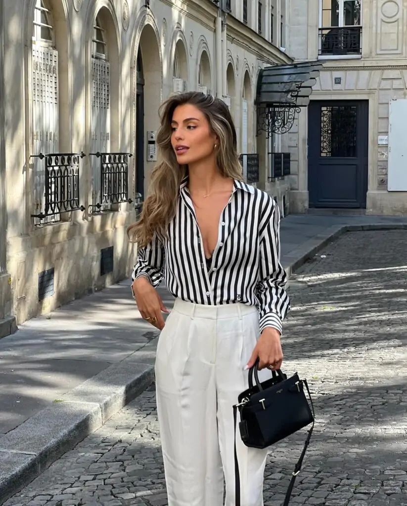 Imagine strolling down a charming European street with elegant architecture. You’re effortlessly chic in a black and white vertically striped shirt, paired with crisp white high-waisted trousers. The look is completed with a small black handbag, adding a touch of sophistication to your ensemble. Perfect for a day of exploration and style.