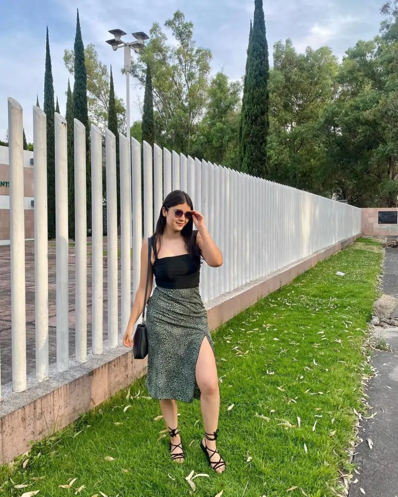 "Step into a serene park and embrace effortless elegance with a stylish twist." You’re rocking a black top paired with a green skirt, featuring a chic slit that adds just the right amount of edge. Black strappy sandals and a sleek black bag complete the look, exuding sophistication. Surrounded by lush greenery and a charming white fence, this ensemble is perfect for a day of relaxed yet fashionable outdoor moments.