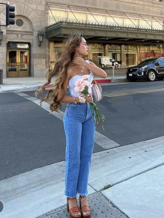 Turn heads this spring with a white strapless top paired perfectly with blue high-waisted jeans. These stylish brown heeled sandals add height and elegance to your ensemble. To top it off, the look is complemented with a fresh bouquet of pink flowers, bringing a vibrant touch to your spring day.