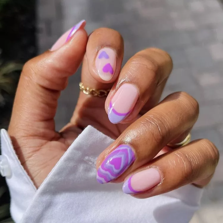 The design is whimsical and fun, perfect for celebrating Valentine's Day with a touch of personality. A playful twist on a classic French manicure features a pastel purple base with a white half-moon design and a swirling heart accent on some nails.