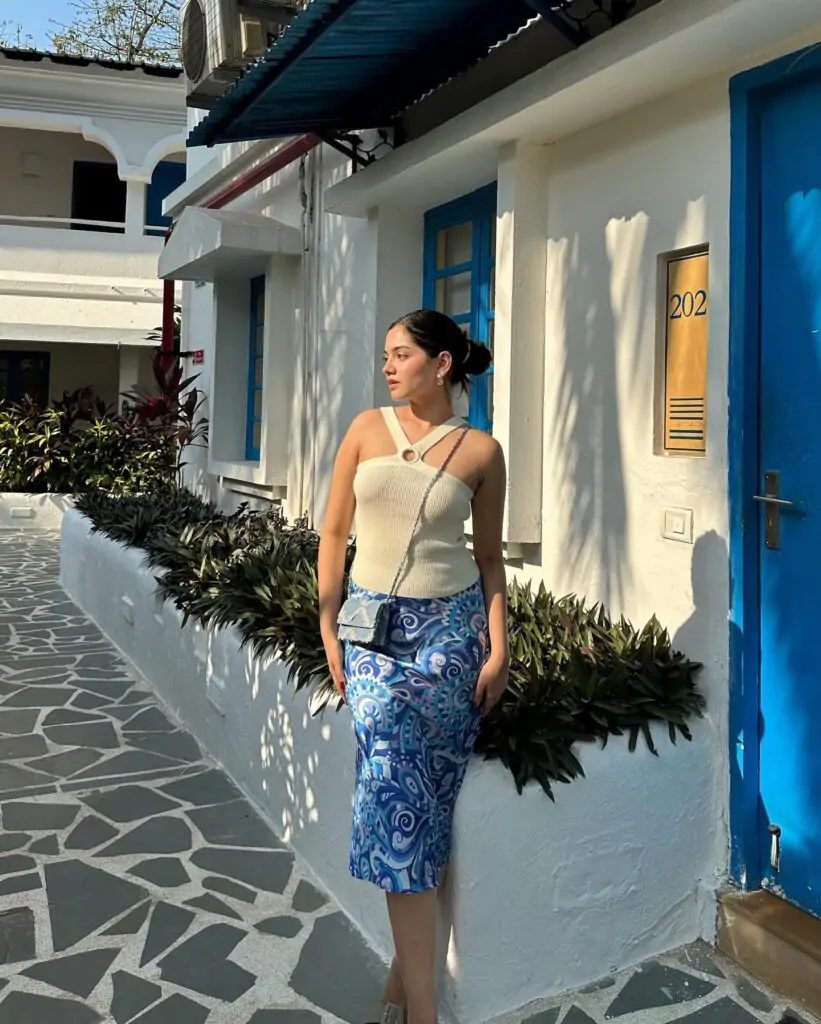 The individual stands confidently in front of a white building with charming blue accents, including a door marked "202." The ground beneath is paved with irregular stone tiles, and lush plants line the wall behind, all basking in the warm sunlight, casting delightful shadows.