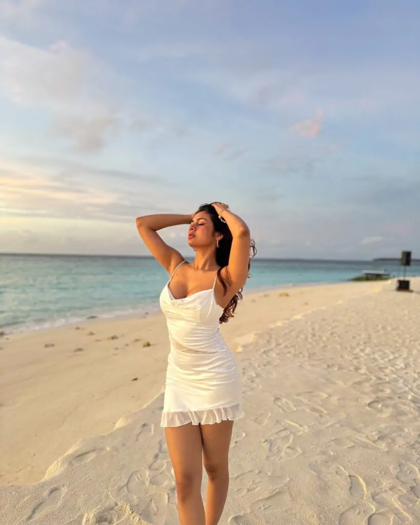 The person stands on a serene beach with gentle waves in the background, and the warm lighting suggests it's either sunrise or sunset.