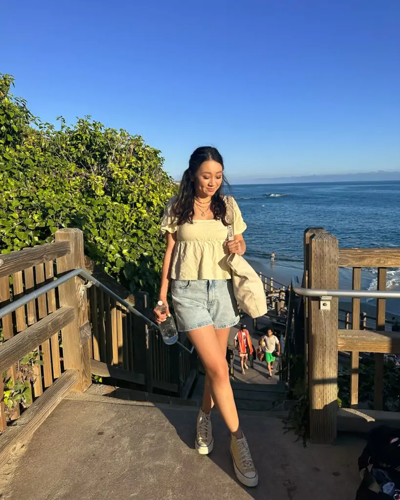 Seaside Elegance: A chic and casual beach outfit featuring a cream peplum top, light denim shorts, and beige high-top sneakers, accessorized with a canvas tote bag and a water bottle, perfect for a relaxed seaside stroll.
