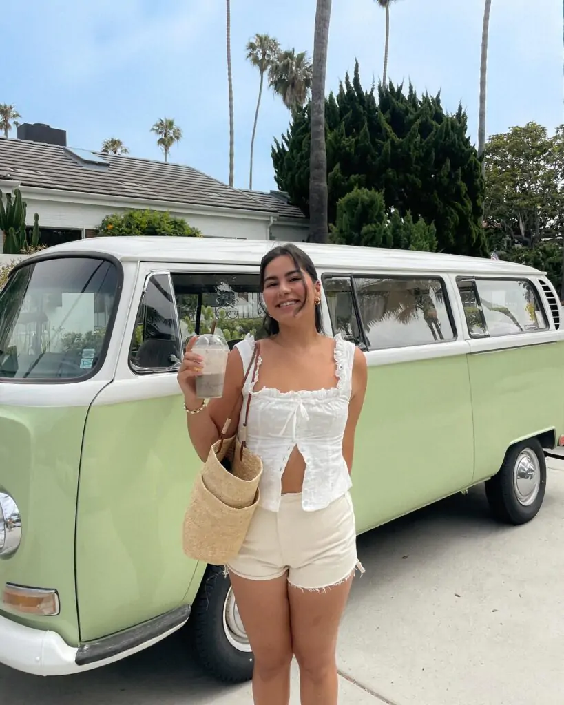 Embrace a retro vibe with a white sleeveless top featuring a chic bow and cut-out detail, light-colored shorts, a straw bag, and fashionable sneakers, all set against a vintage Volkswagen van backdrop.