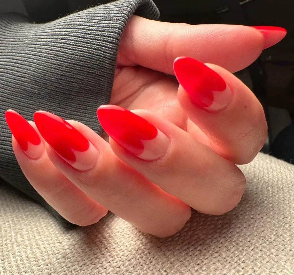 This Valentine's Day manicure is a bold and romantic take on the classic French tip. Each nail is painted a vibrant red with a subtle heart shape cut out at the tip, showcasing a sheer, nude base underneath.