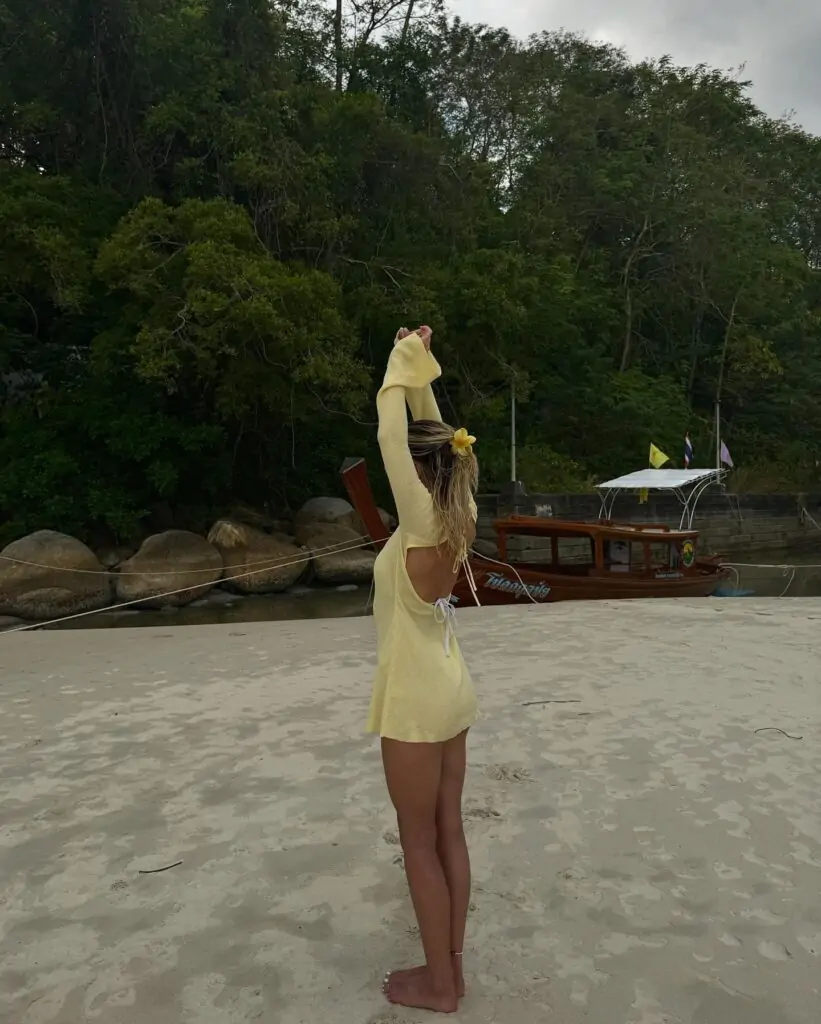 Stay stylish by the shore—“Seaside Elegance” features a light yellow open-back dress, adorned with a yellow flower in the hair, perfect for a serene beach day amidst lush greenery and rocky shores.