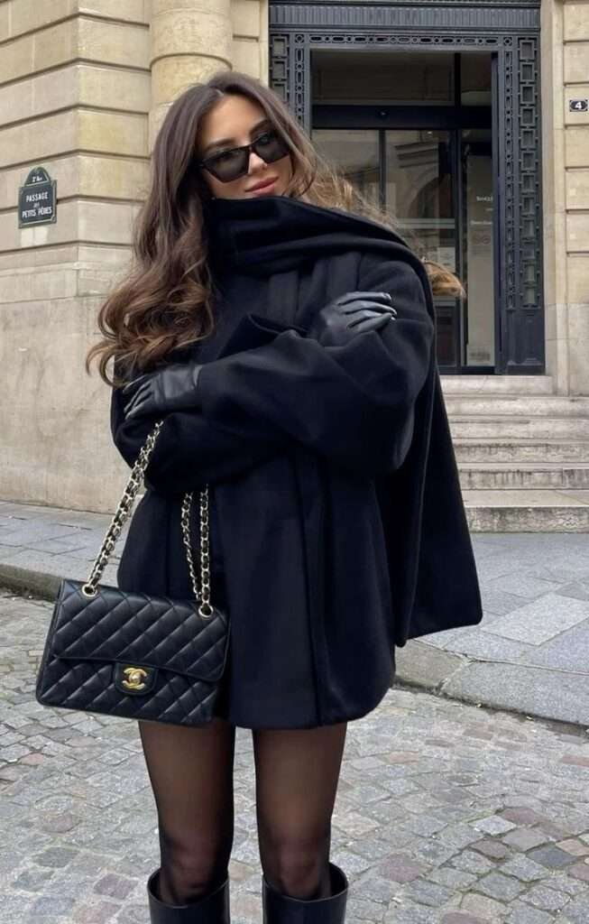 Mystery in Monochrome: The look centers on a dramatic, sweeping black wool cape or cocoon coat with an integrated high collar, channeling effortless, old-school Parisian glamour. Essential Accessories: Sharp leather gloves and opaque black tights emphasize the sophisticated all-black foundation, while sleek sunglasses maintain an air of powerful anonymity. The Ultimate Classic: The outfit is instantly elevated to icon status with the timeless addition of a quilted black leather shoulder bag with a gold chain strap.