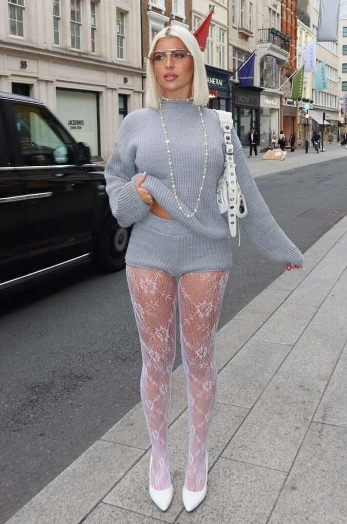 This image captures a playful, directional streetwear moment, which I would call "Lace & Luxe Knitwear".
🧶 Lace & Luxe Knitwear
The ensemble is built on striking texture contrast, pairing a cozy, monochromatic gray ribbed knit set—featuring a slightly cropped mock-neck sweater and matching shorts—with a provocative element. This foundation is dramatically offset by elaborate white floral lace fishnet tights and sharp white pointed-toe pumps, creating a high-impact tension between soft comfort and bold femininity, completed with oversized silver accessories and modern sunglasses.