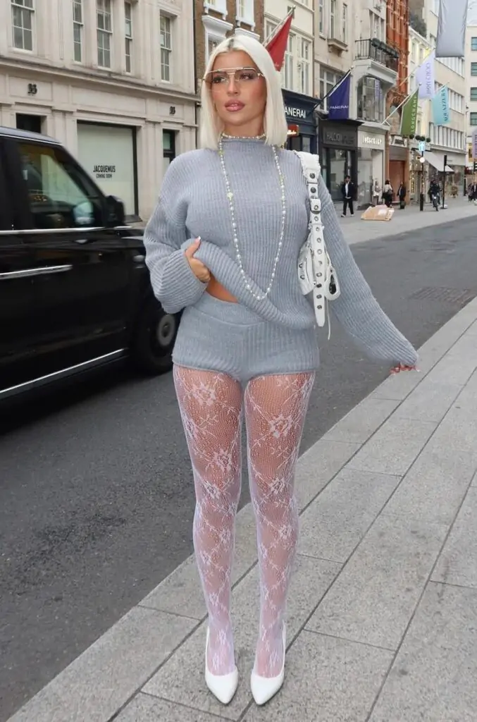 
This image captures a playful, directional streetwear moment, which I would call "Lace & Luxe Knitwear".

🧶 Lace & Luxe Knitwear
The ensemble is built on striking texture contrast, pairing a cozy, monochromatic gray ribbed knit set—featuring a slightly cropped mock-neck sweater and matching shorts—with a provocative element. This foundation is dramatically offset by elaborate white floral lace fishnet tights and sharp white pointed-toe pumps, creating a high-impact tension between soft comfort and bold femininity, completed with oversized silver accessories and modern sunglasses.