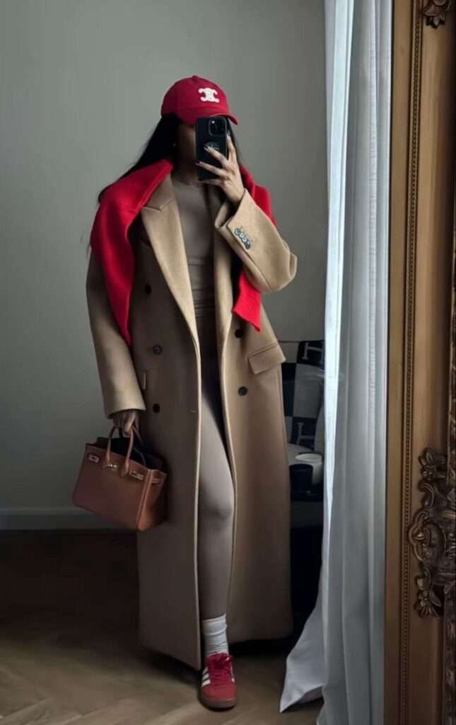 Master the art of high/low dressing by pairing a floor-sweeping, impeccably tailored camel coat with a sleek, monochromatic nude activewear set underneath. The power is in the accessories: a vibrant, fire-engine red baseball cap and matching scarf inject an essential, playful sports luxe energy. Finished with quiet luxury via a structured Birkin-style leather tote, this look perfectly blends cozy comfort with sophisticated, dynamic style.