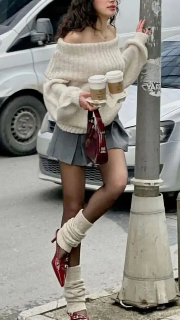 Texture Play: This look pairs a voluminous, soft off-the-shoulder knit with a structured micro-pleated gray skirt, striking the perfect balance between cozy and chic. Embrace the contrast: Sheer black tights meet slouchy ivory leg warmers, creating a playful, layered aesthetic that leans into the trending balletcore movement. The essential pop: Finish with high-shine patent red Mary Janes and a matching miniature handbag, ensuring every step makes a dramatic, unforgettable statement.